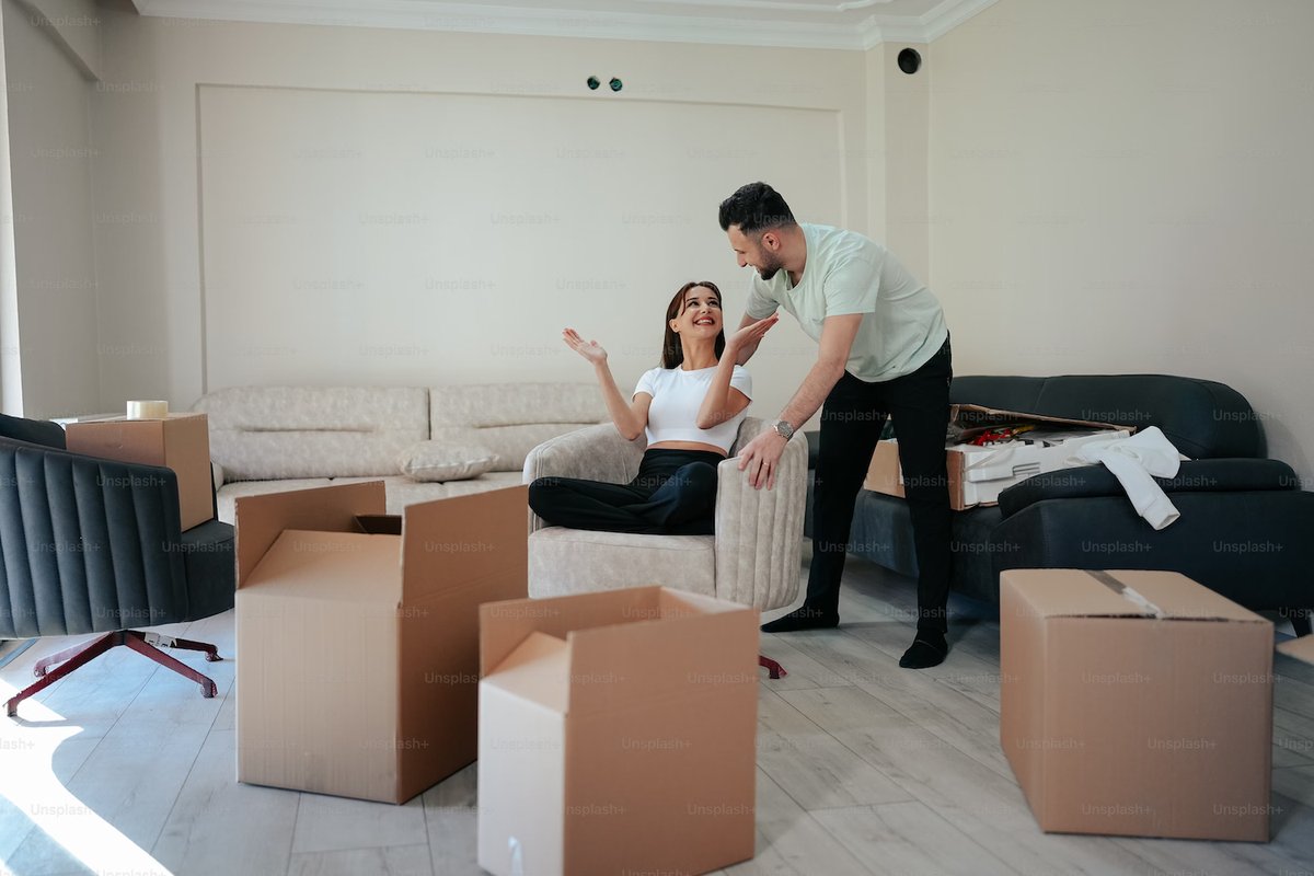 Smiling woman sitting cross-legged in an armchair while a man leans in to talk to her in a room with moving boxes.