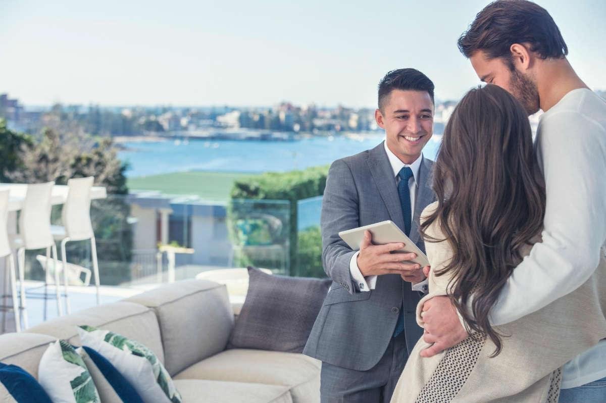 Smiling real estate agent showing a tablet to a couple on a balcony with a waterfront view.